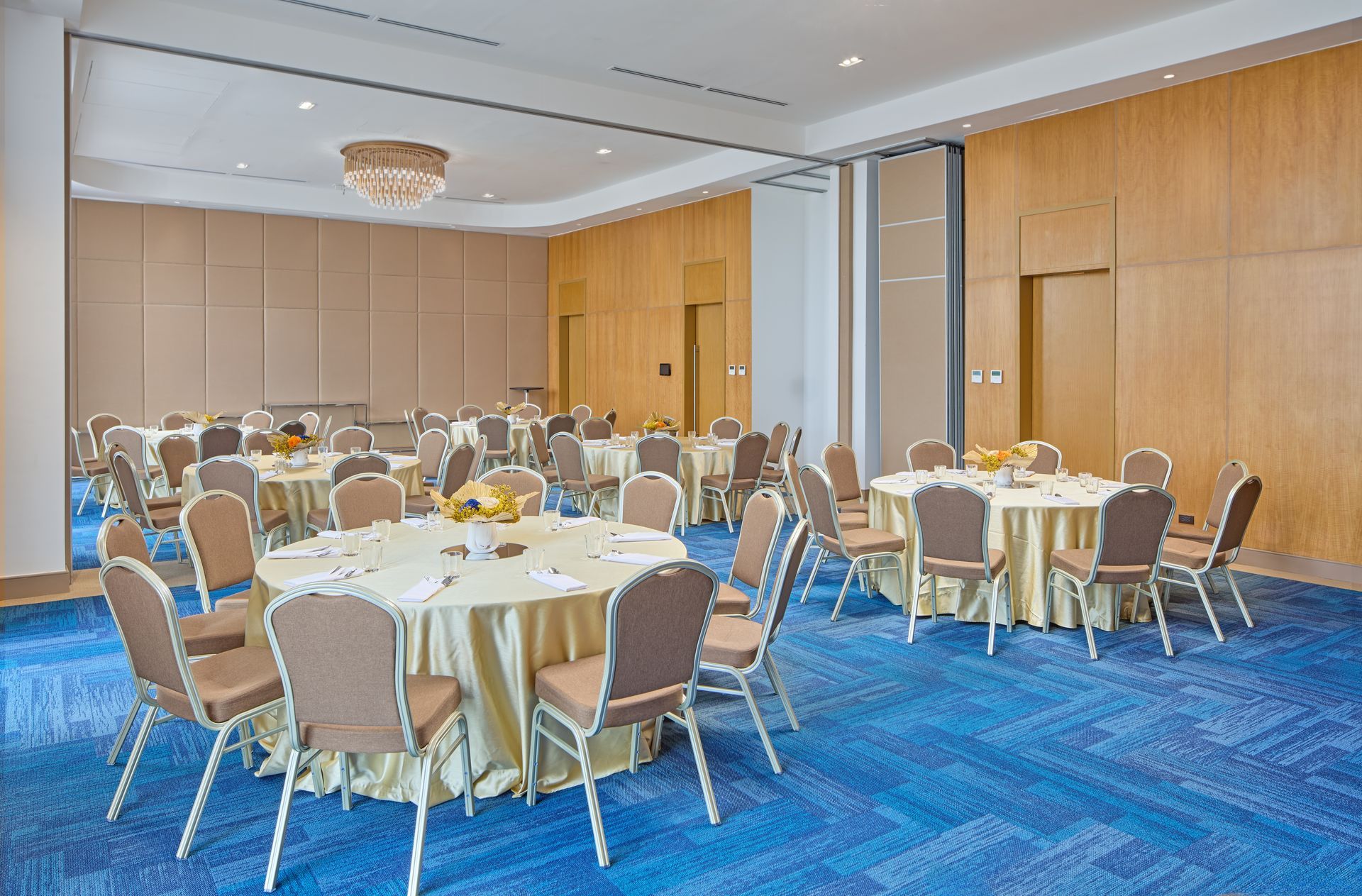 A banquet hall with round tables covered in beige cloths, surrounded by chairs on a patterned blue carpet.
