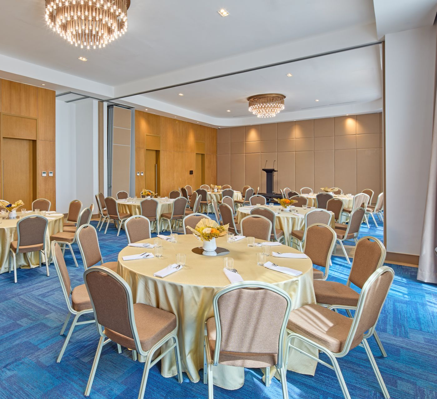 A banquet hall with round tables covered in beige tablecloths, surrounding chairs, and two ornate crystal chandeliers.