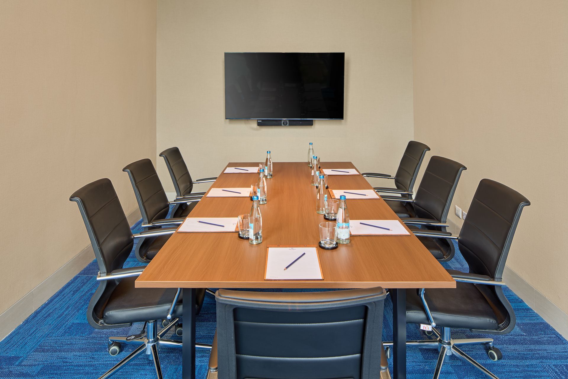 A small meeting room featuring a wooden table surrounded by eight black chairs, with a television mounted on the wall.