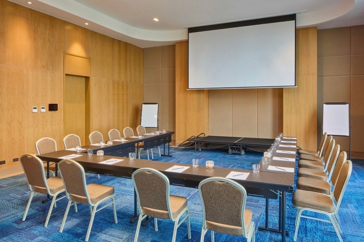 A modern conference room with rows of tables and chairs, wood-paneled walls, a podium, and large windows with blue carpet.
