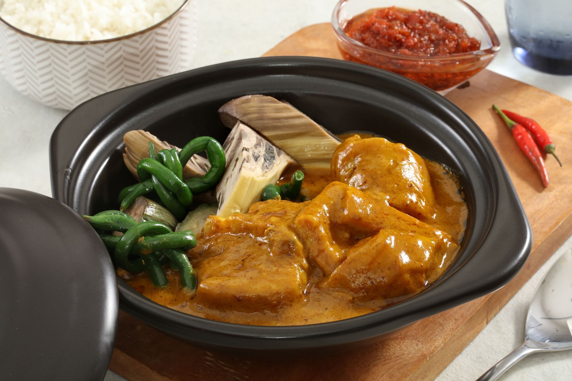 A bowl of shrimp sinigang with vegetables, served with a side of rice, dipping sauce, and chili peppers on a wooden board.