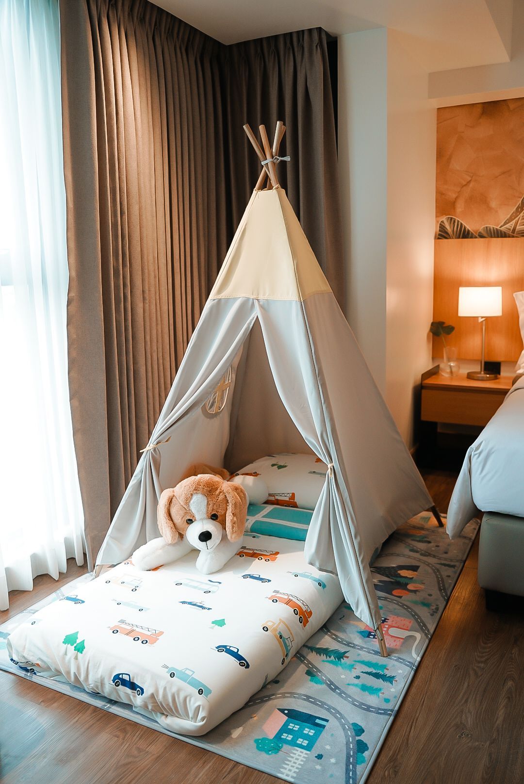 A children's teepee tent with a stuffed dog inside, on a colorful play mat next to a hotel bed.