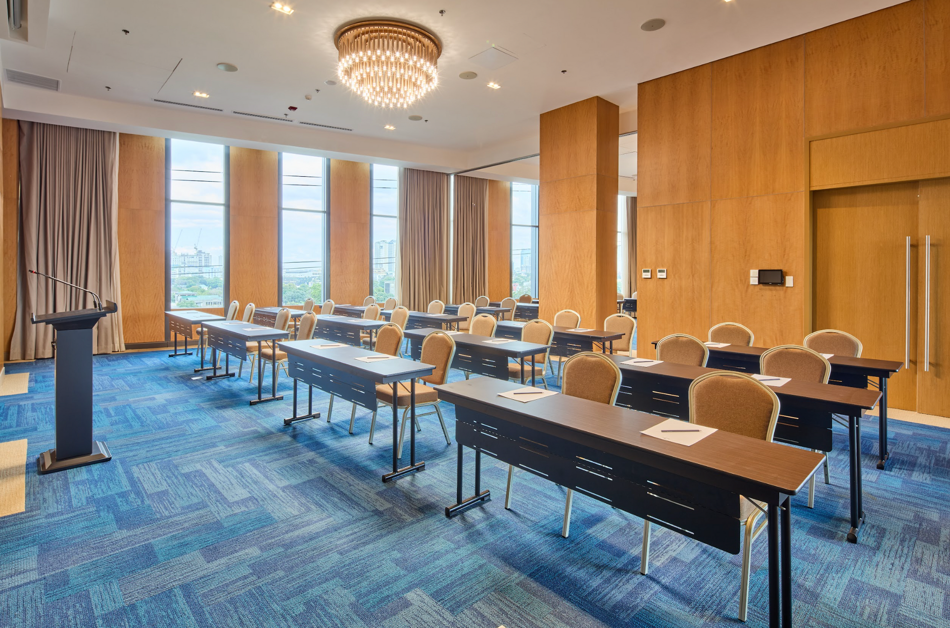 A well-lit conference room with rows of tables and chairs, wood-paneled walls, a blue patterned carpet, and chandeliers.