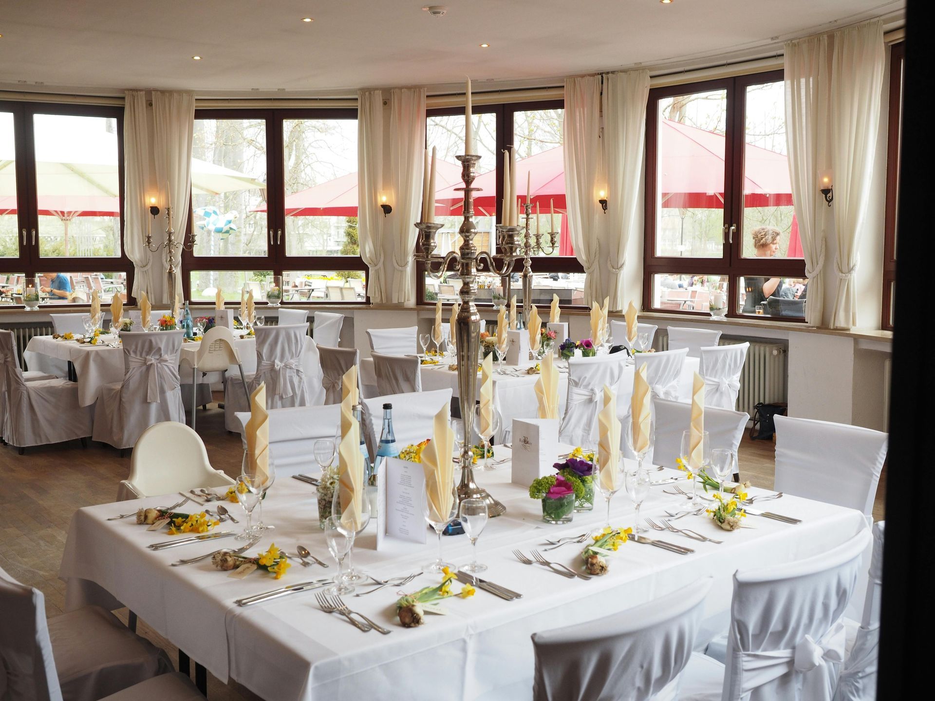 Formal dining room set for an event with white tablecloths, chairs, and centerpieces with candles, and outdoor views.
