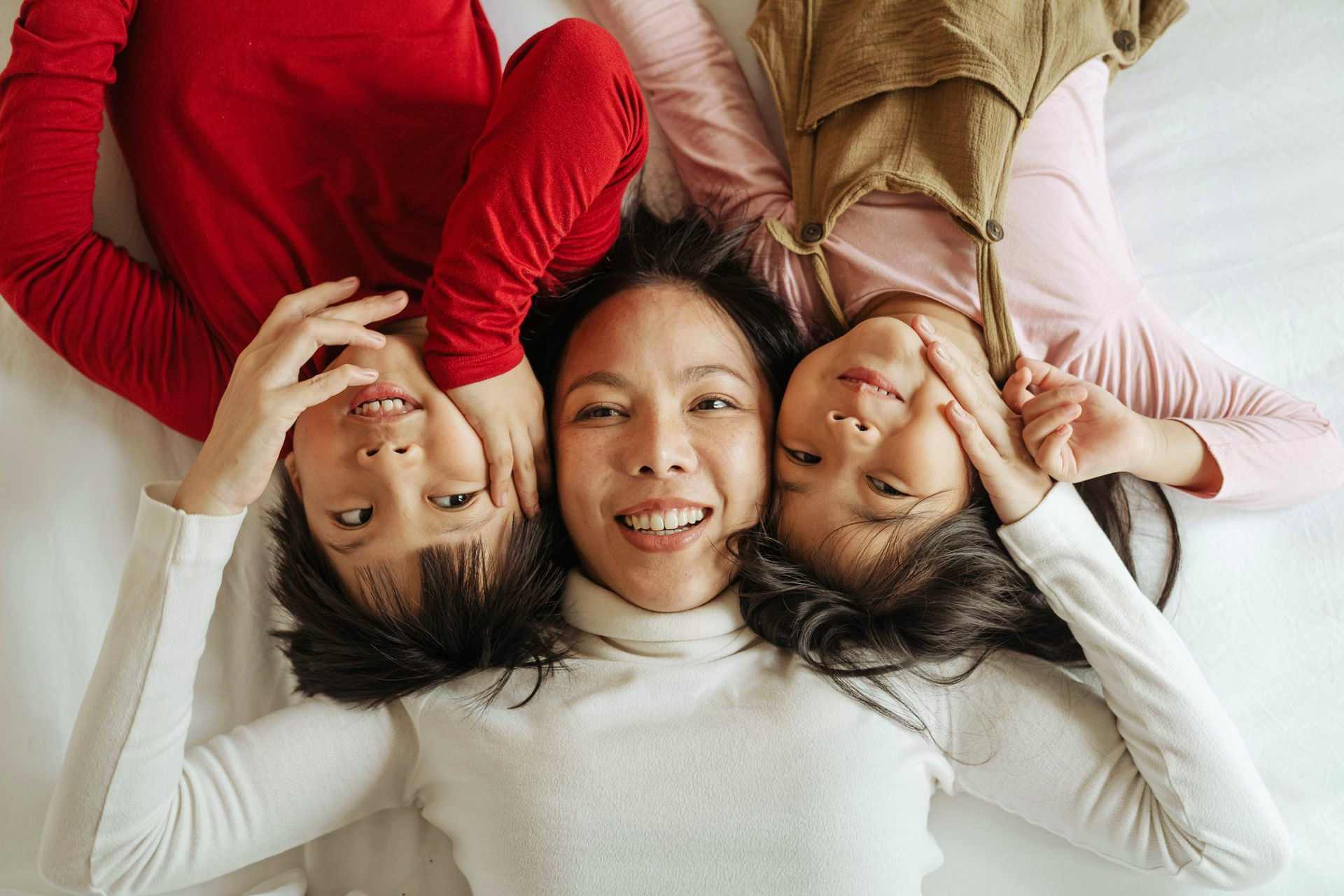 A woman and two children lie side-by-side on a white surface, smiling while touching their cheeks.