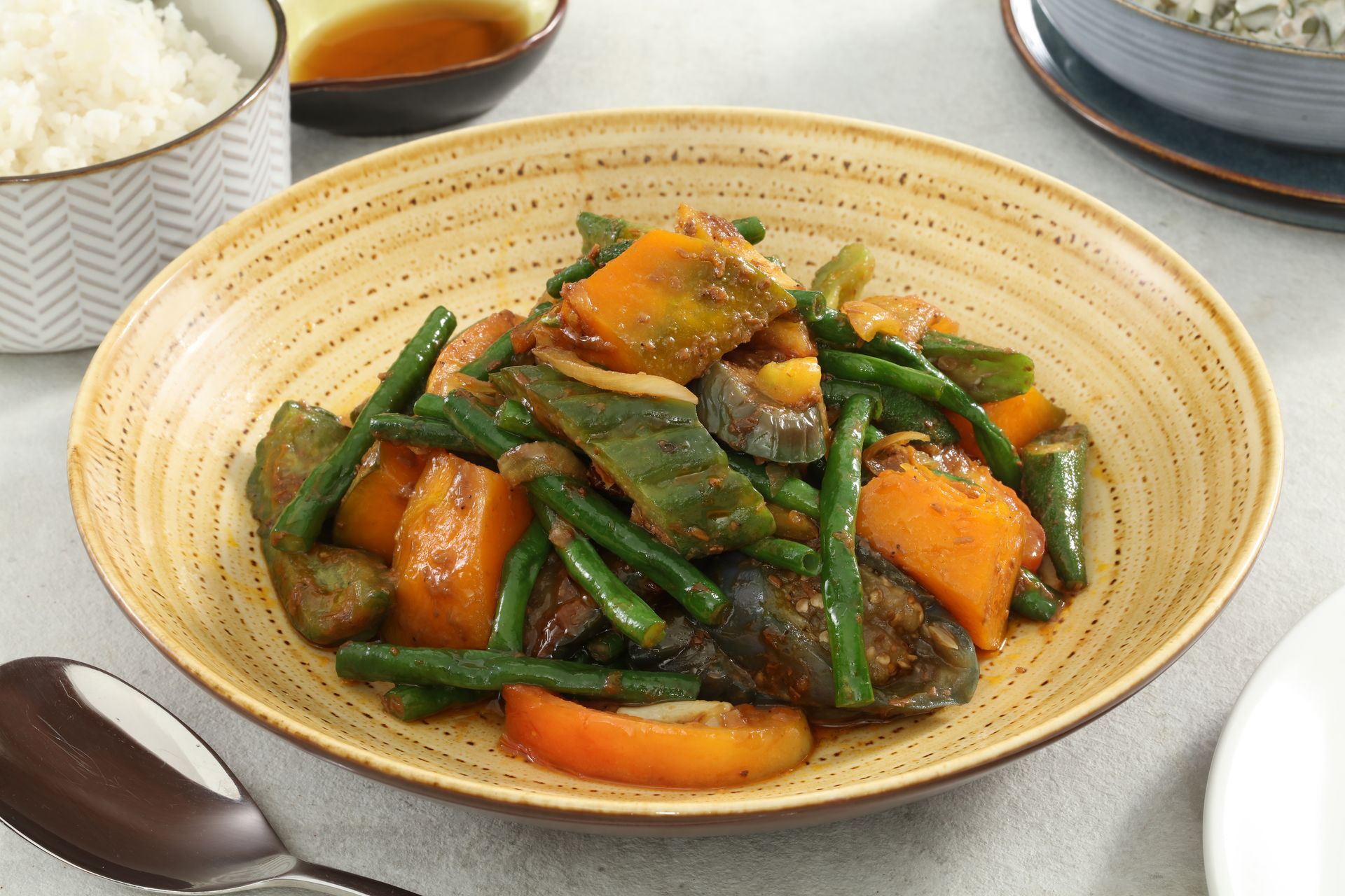 A bowl of vegetable stir-fry with green beans, squash, and eggplant, served next to a small bowl of rice and dipping sauce.