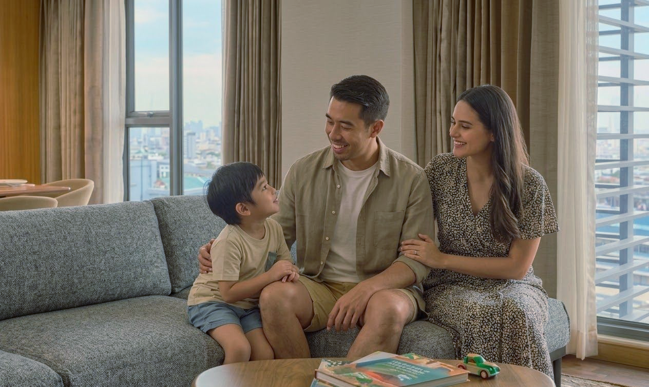 A family sits together on a gray couch in a bright living room, smiling at each other by a window overlooking a city.