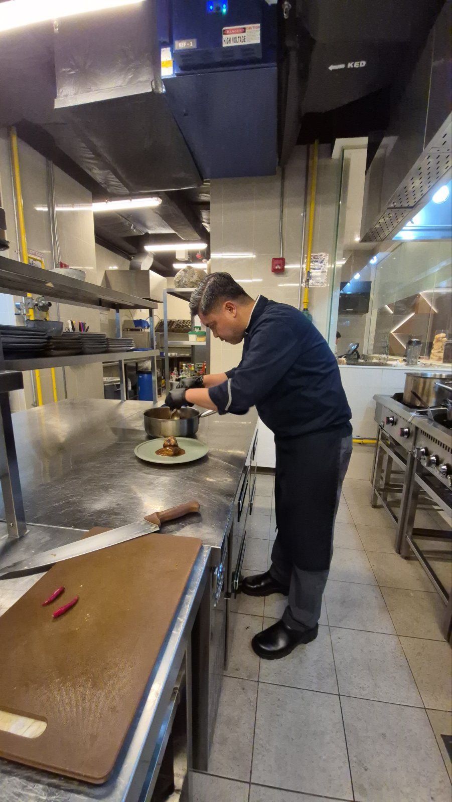 Person plating food in a commercial kitchen, wearing dark clothes and working at a stainless steel counter.