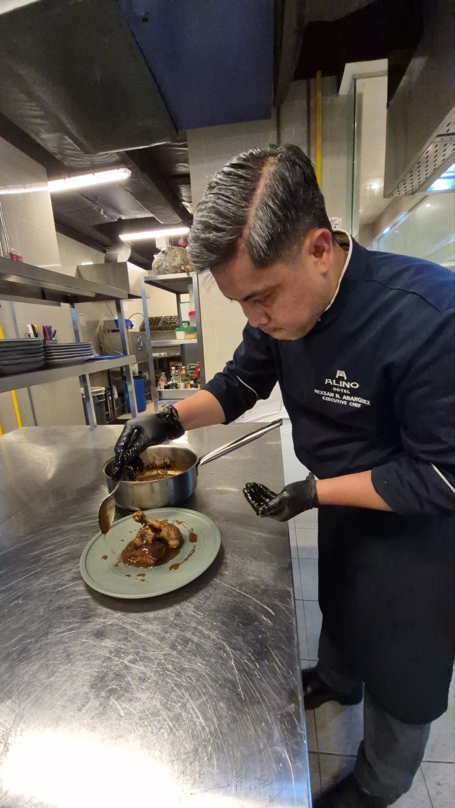 Chef plating food in a stainless-steel kitchen, focused over a dish on the counter.