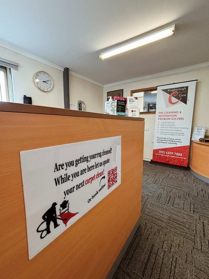 A sign on a wooden reception desk promotes carpet cleaning services, with a QR code and a banner stand in the background.