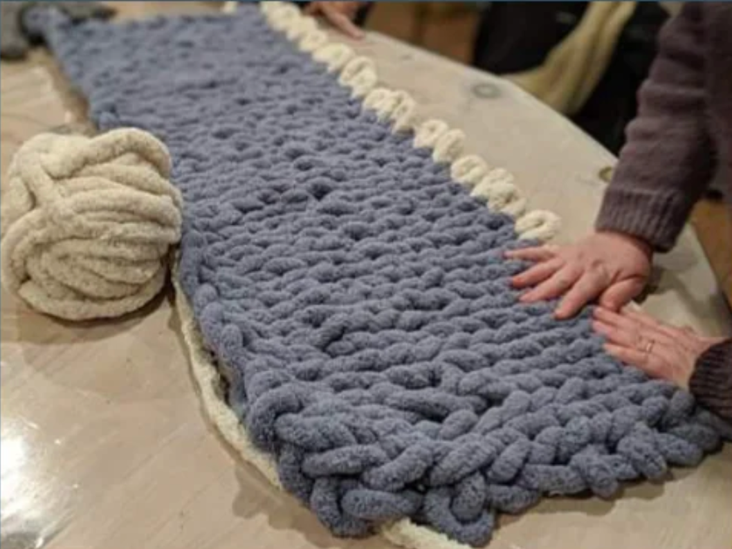 A person's hands smoothing out a large, chunky knit blanket on a table next to a ball of yarn. The blanket is blue with a cream-colored border.