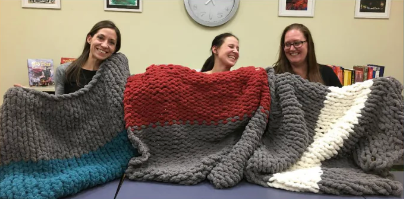 Three people holding up large, colorful knitted blankets.