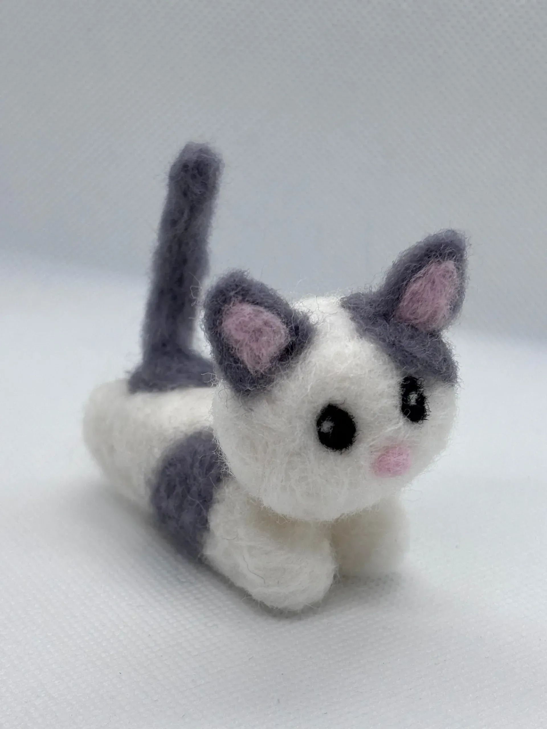 A small, handmade felt cat figure with white fur, grey patches, a grey tail, and a tiny pink nose on a white background.