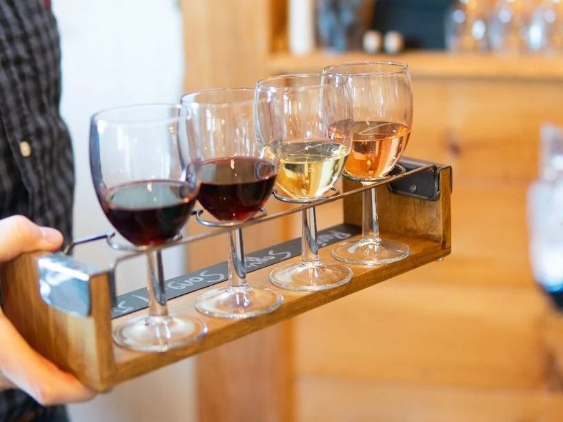 A person is holding a wooden tray with four glasses of wine on it.
