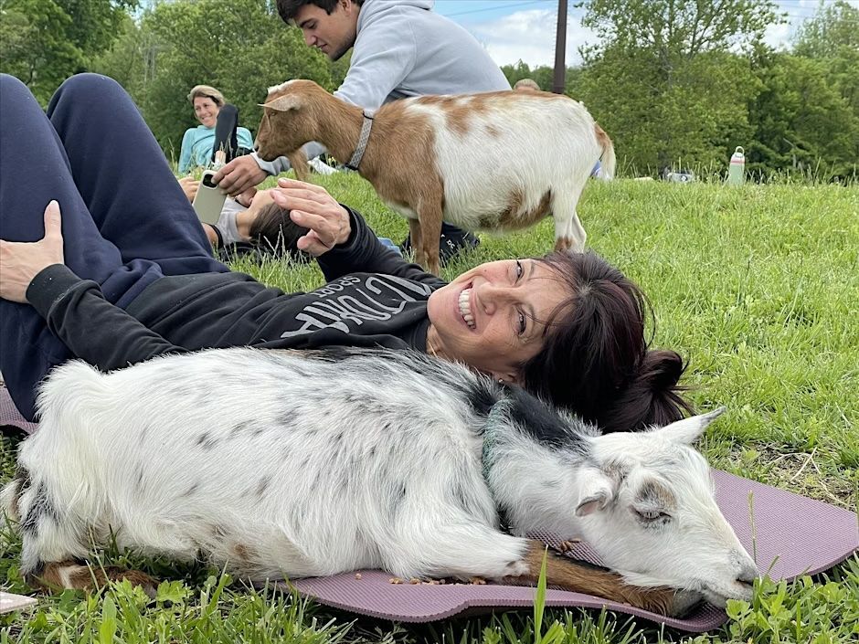 Woman smiles, lying on a yoga mat with a goat, while another goat and person stand nearby in a grassy field.