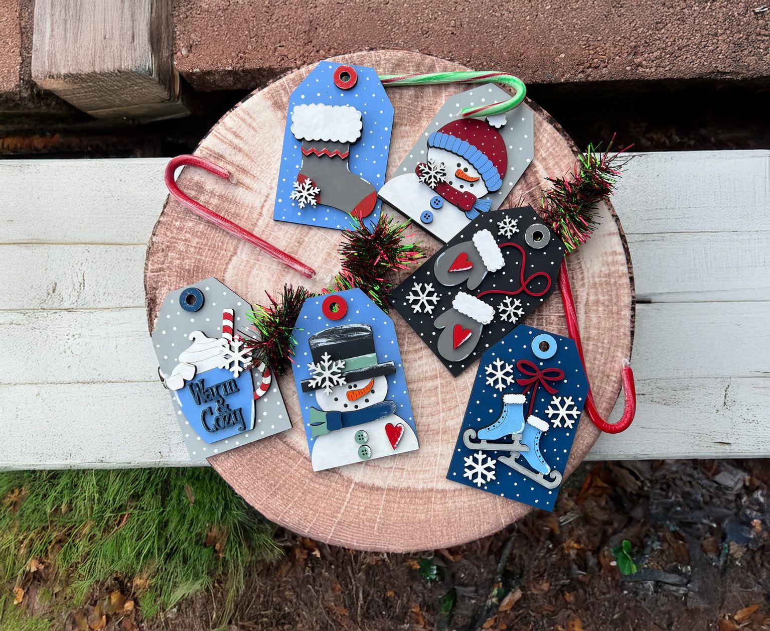 Christmas gift tags on a wood slice: snowman, stocking, hot cocoa, ice skates, mittens, snowflakes; red, blue, white, black.
