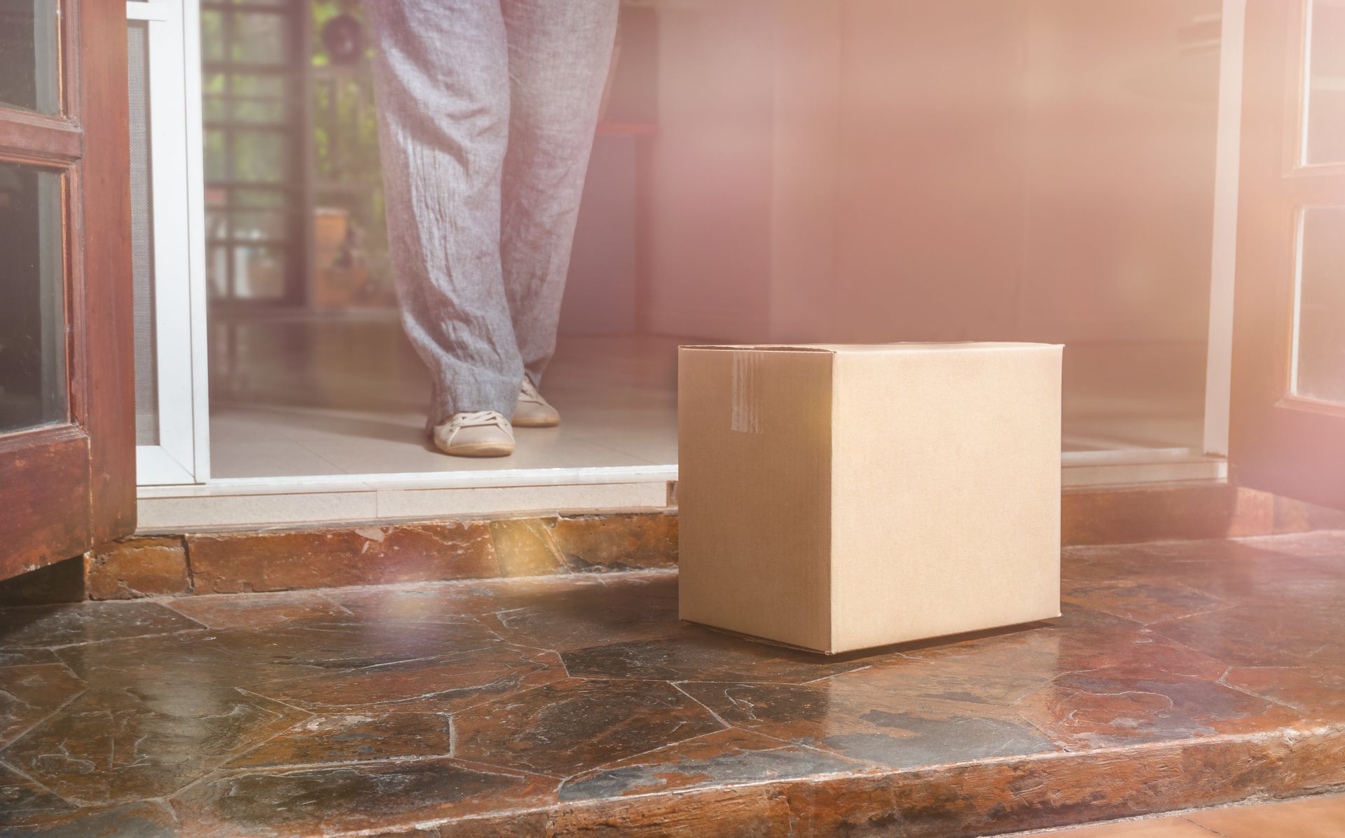 Cardboard box on a doorstep, near legs and feet of a person inside a doorway.