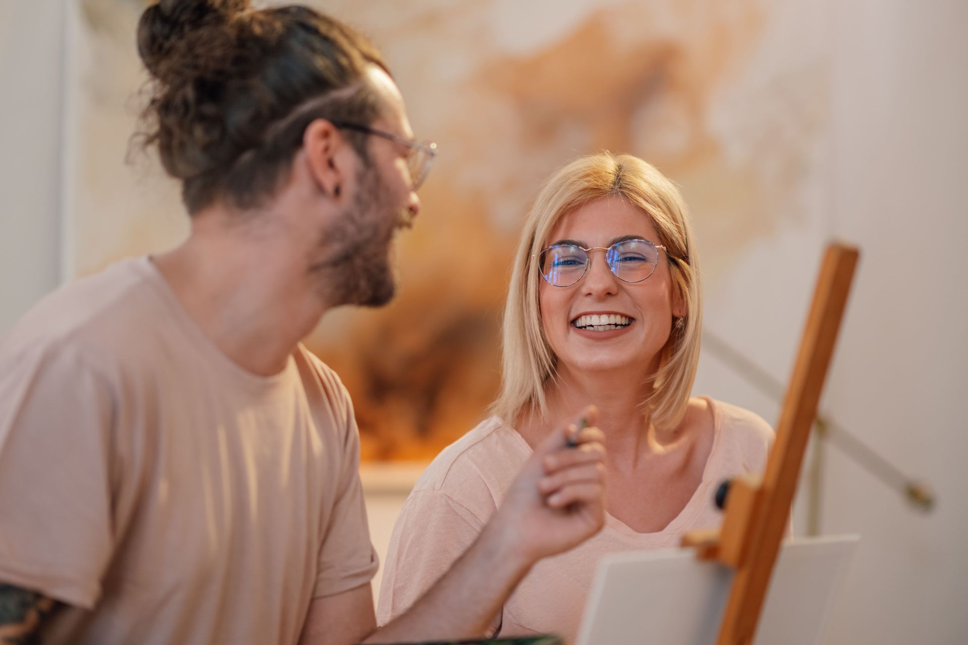 Man and woman smiling, sitting near an easel with a painting, likely indoors.