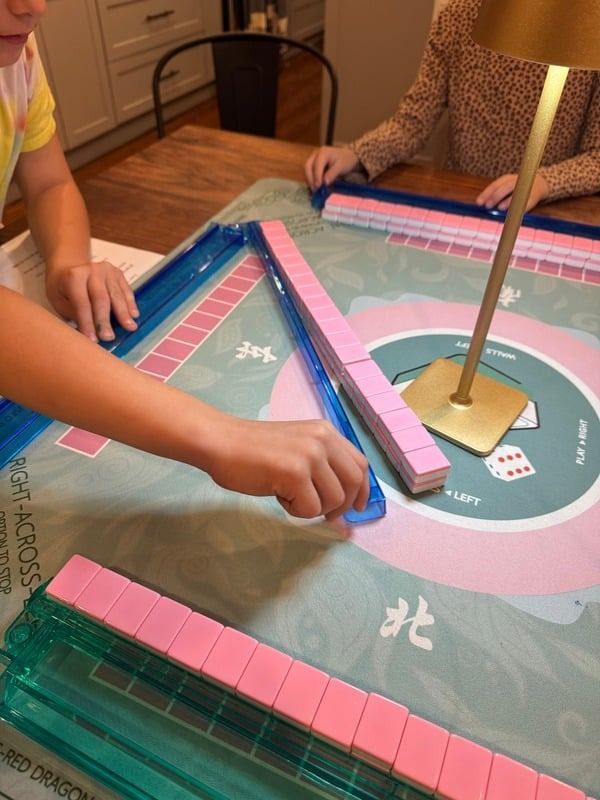 Two people sit at a table playing a mahjong game with pink tiles arranged on a specialized mat.