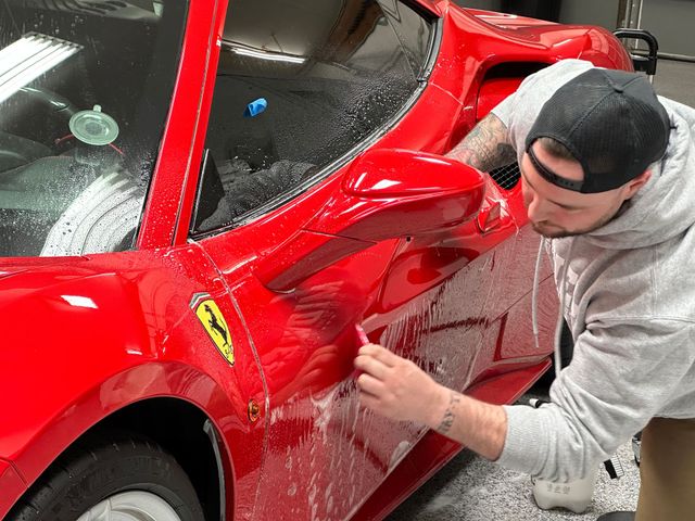 Man polishing a red Ferrari in a garage, wearing a gray hoodie and black cap.