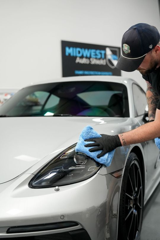 Detailing a silver sports car’s hood with a blue cloth in a garage, with a Midwest Pro Detailing sign in the background