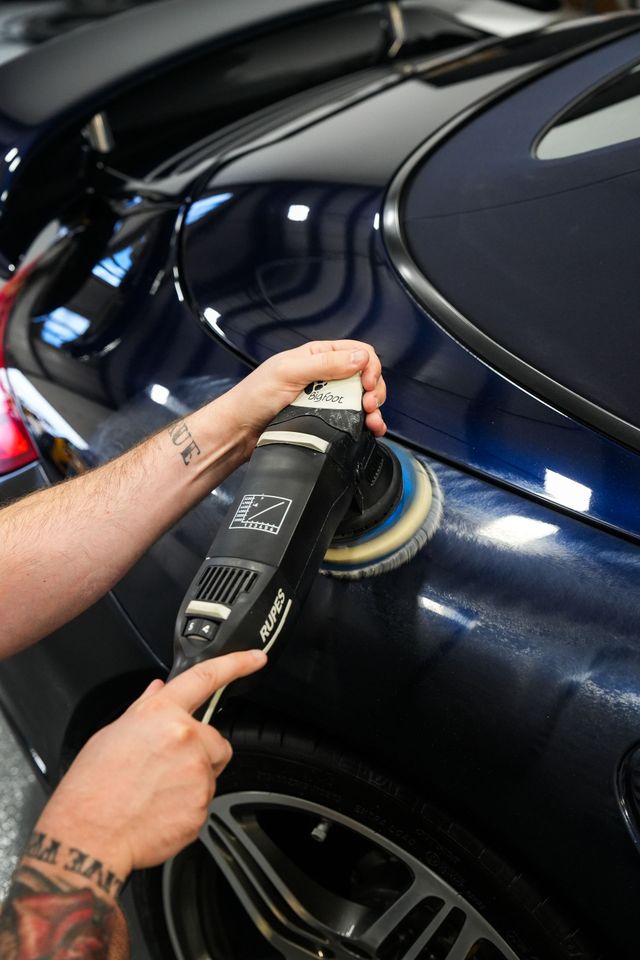 Hand polishing a dark blue car with a power buffer in a garage