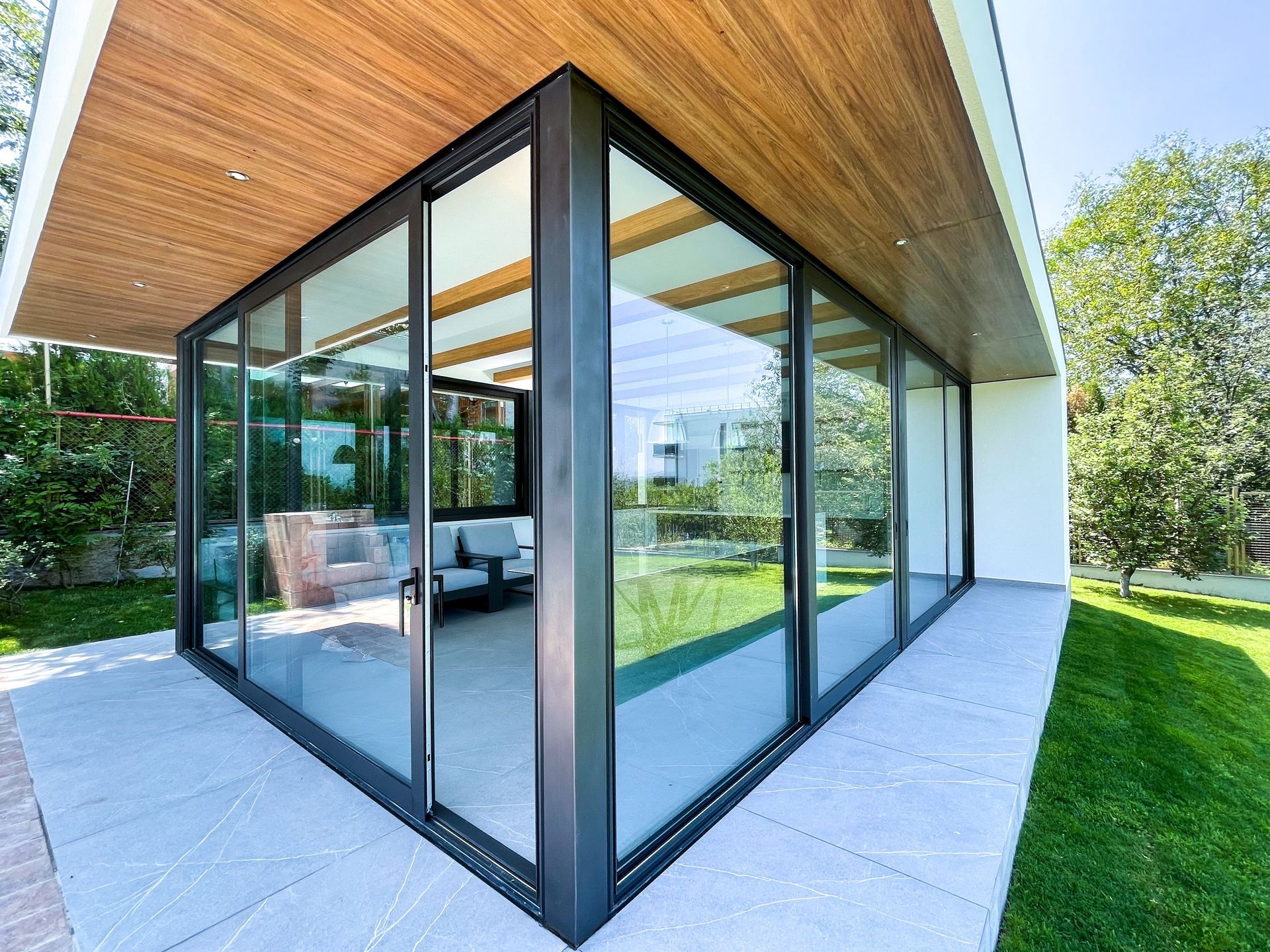 Modern glass-walled patio room with wood سقiling, opening to a green lawn and garden