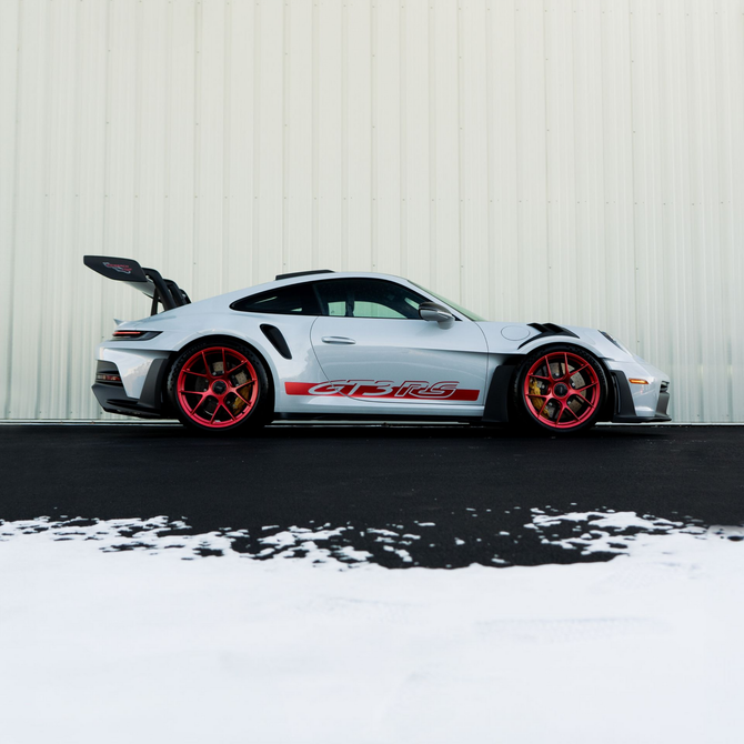 White sports car with red rims and accents parked on snow beside a corrugated wall.
