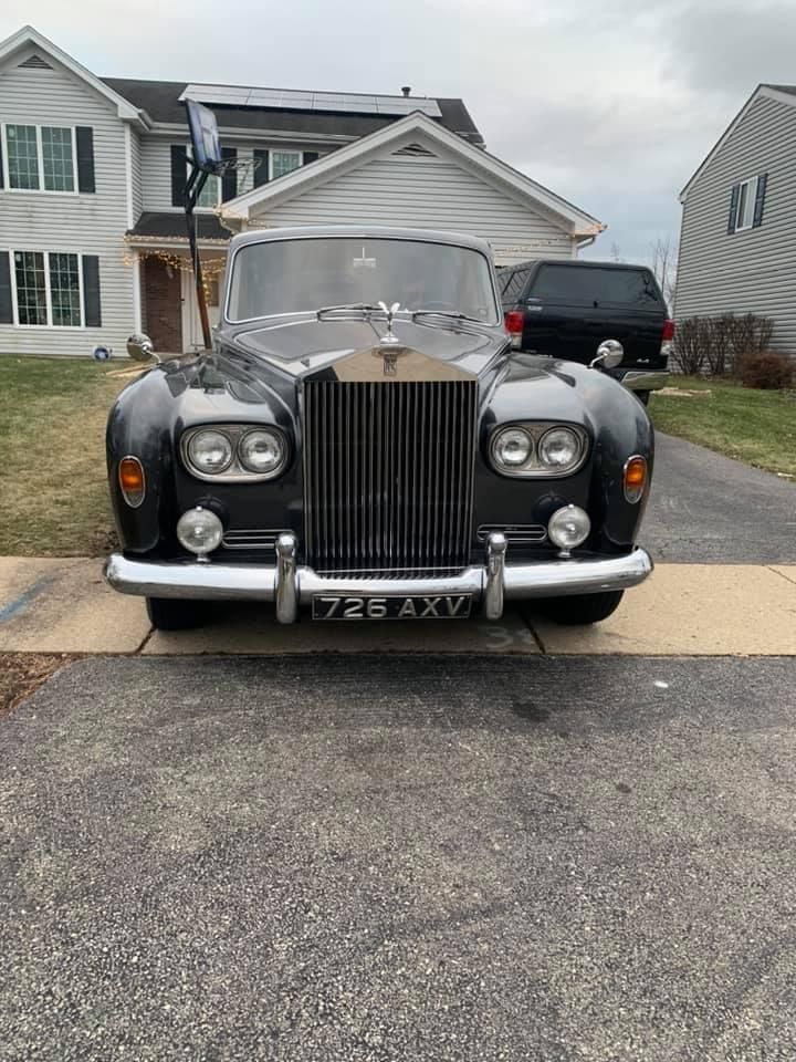 Classic black Rolls-Royce parked in a suburban driveway, front hood open, with houses in the background.