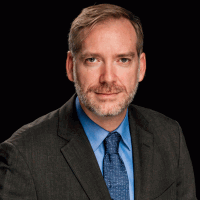 Man in suit, blue shirt, and tie. Dark background, looking at camera, neutral expression.