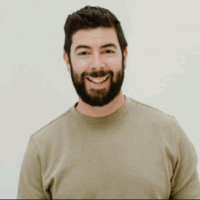 Man with beard smiling, wearing a light green sweater, standing in front of a white wall.