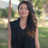 Woman with long dark hair smiles, wearing a black vest and shirt, in a park setting.