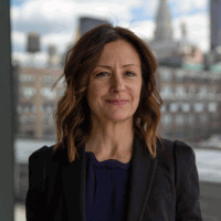 Woman with brown hair, wearing a black blazer, smiling, cityscape background.