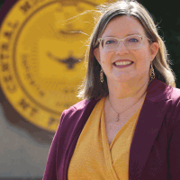 Woman in glasses and a purple blazer smiles in front of a Central Michigan University logo.