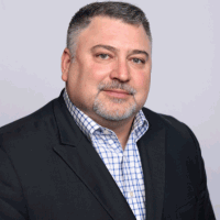 Man in suit jacket, smiling, looking directly at the camera against a light gray background.