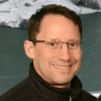 Man with glasses smiling, wearing black jacket, in front of a blurred, natural background.