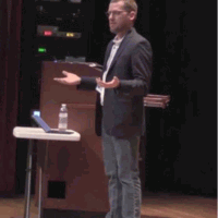 Man gesturing during a presentation, with laptop, water, and books on a table. He is standing in front of a dark stage.