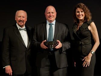 Three people on stage, award recipient holding trophy; two others flank him.