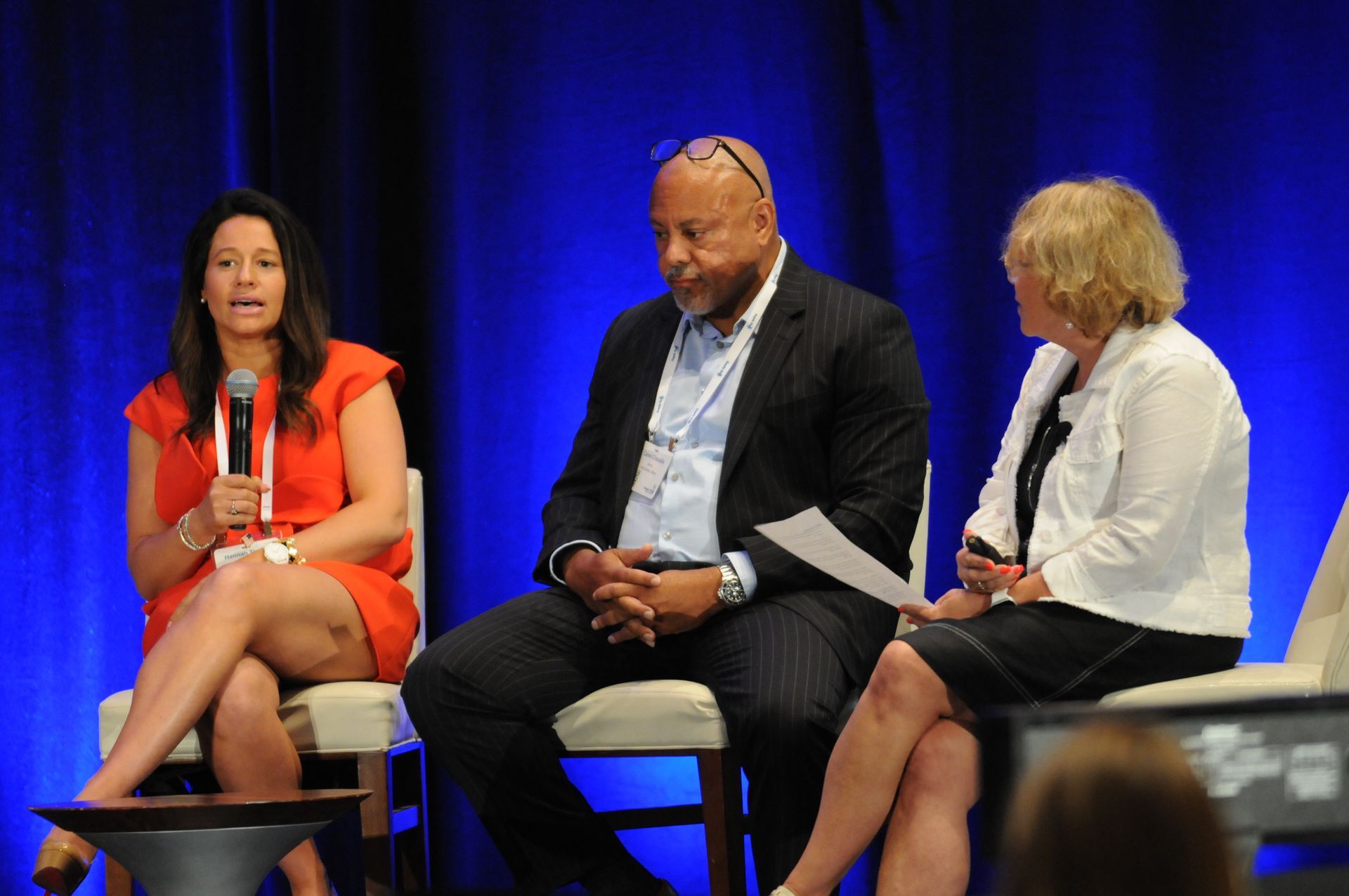 Panel of three people on stage, woman in red dress speaking, man in suit, woman in white jacket holding paper.