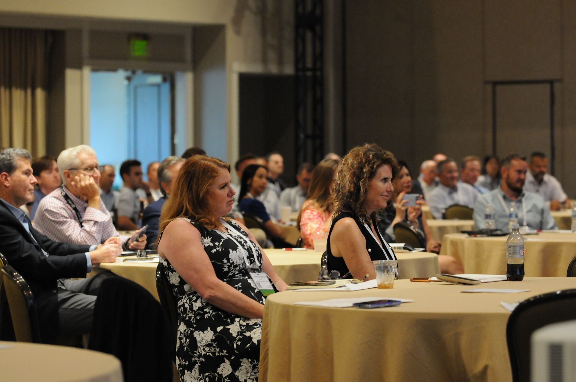 Audience at a conference listening to a presentation. People seated at round tables in a large room.