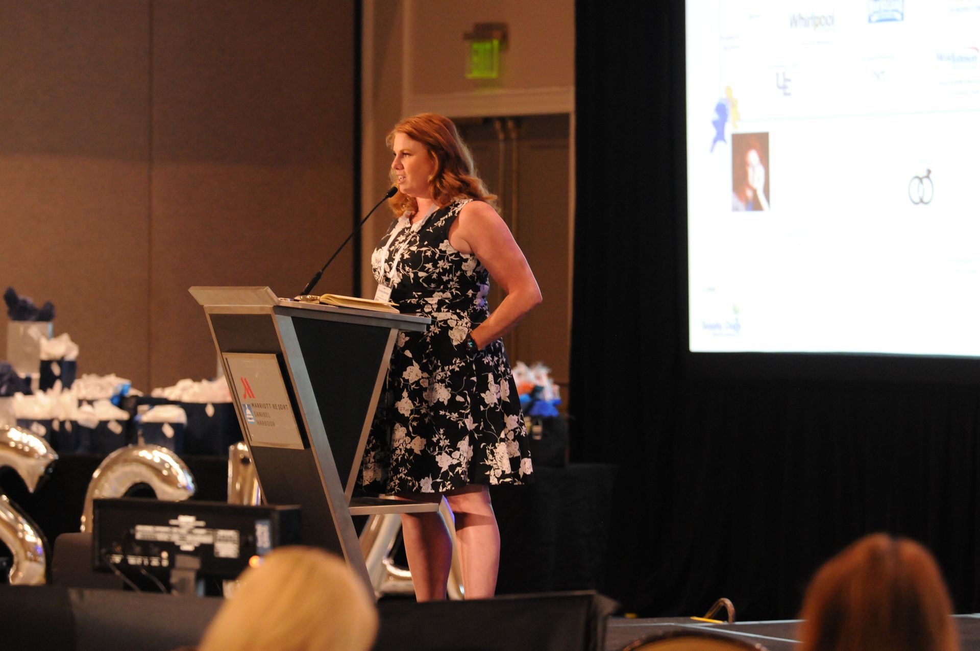 Woman speaking at a podium on stage, wearing a black and white dress, with a screen and balloons in the background.