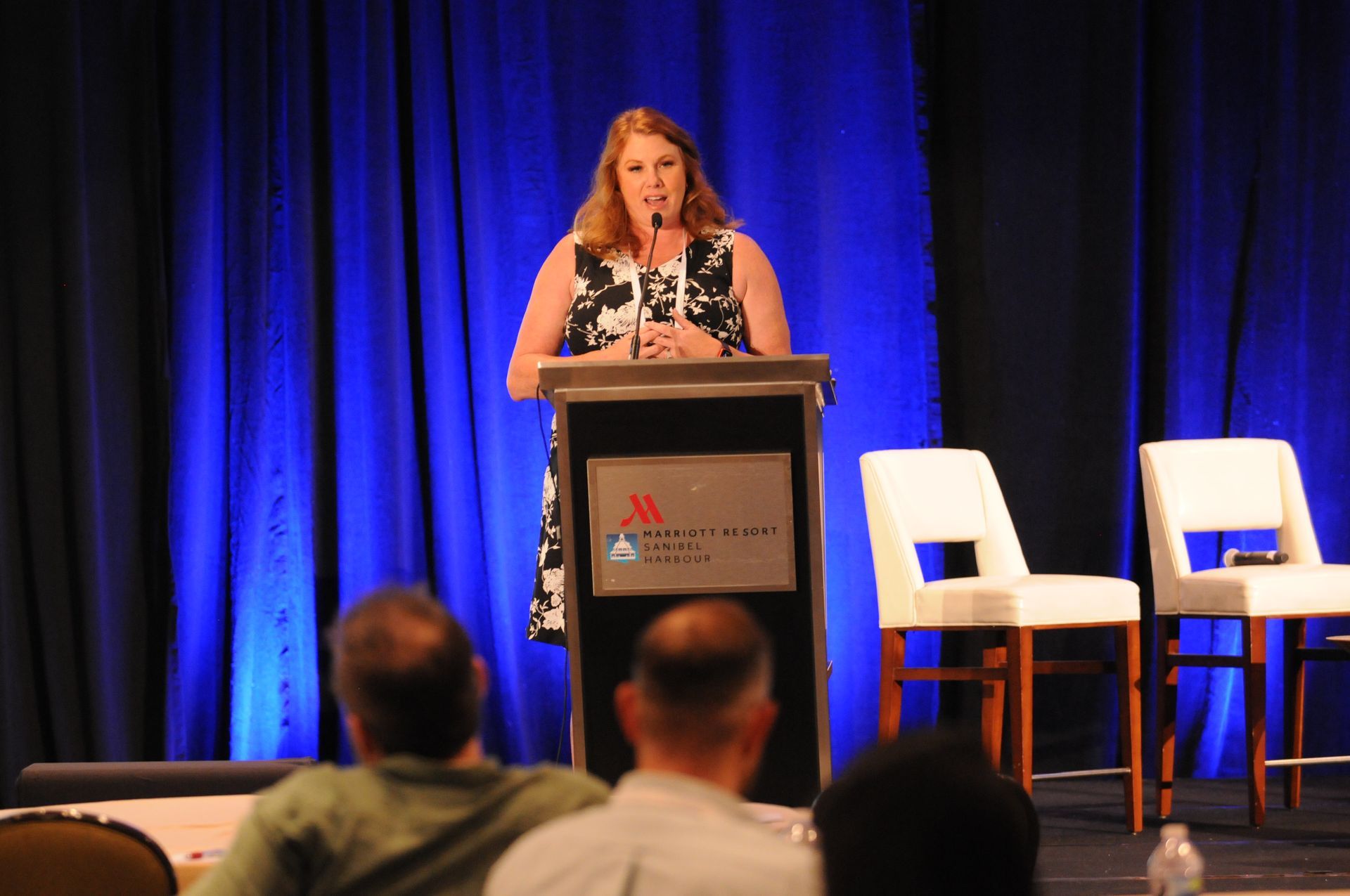 Woman speaking at a conference. She stands at a podium with a black and white dress, in front of a blue curtain.