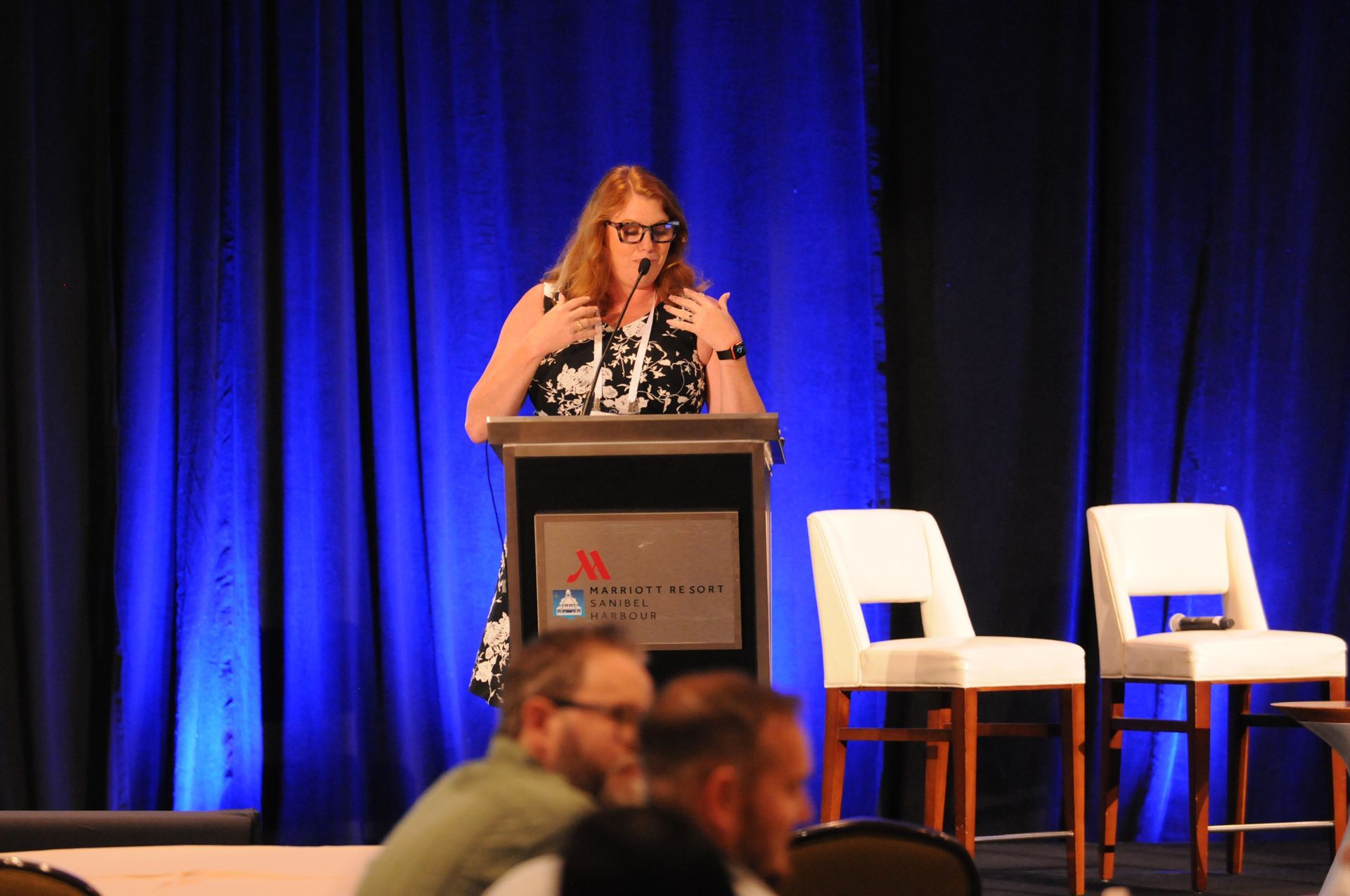 Woman giving a presentation at a Marriott podium, blue curtains behind her, white chairs on either side.