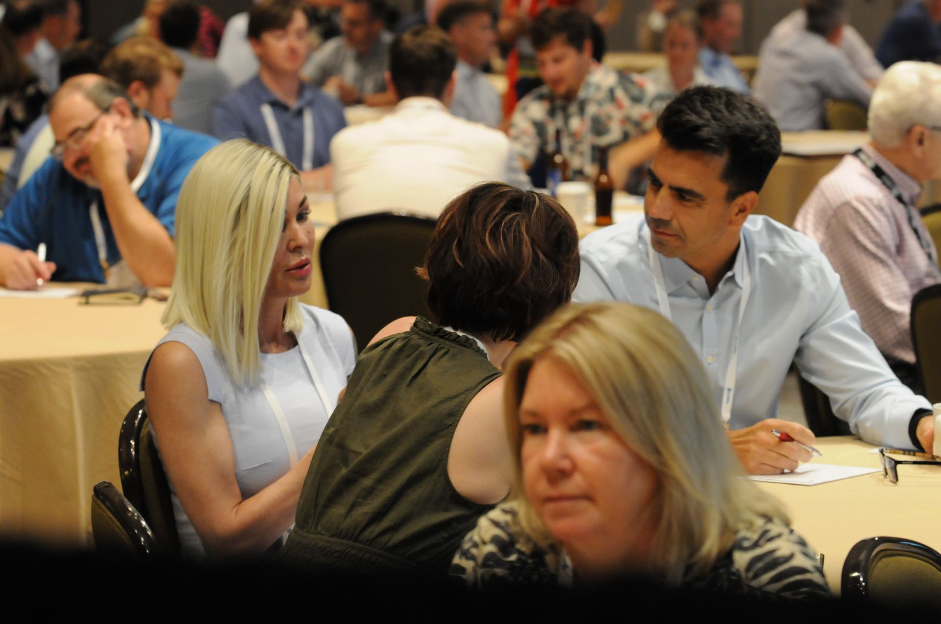 People at a conference, some are talking, others taking notes. Tables, chairs, and diverse attendees.