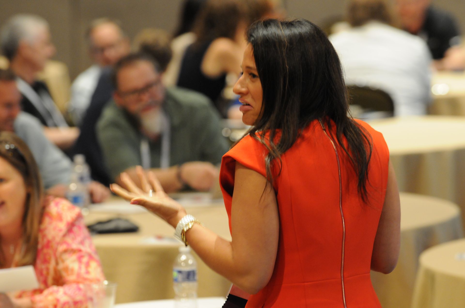 Woman in orange dress gestures while speaking to audience in a conference setting.