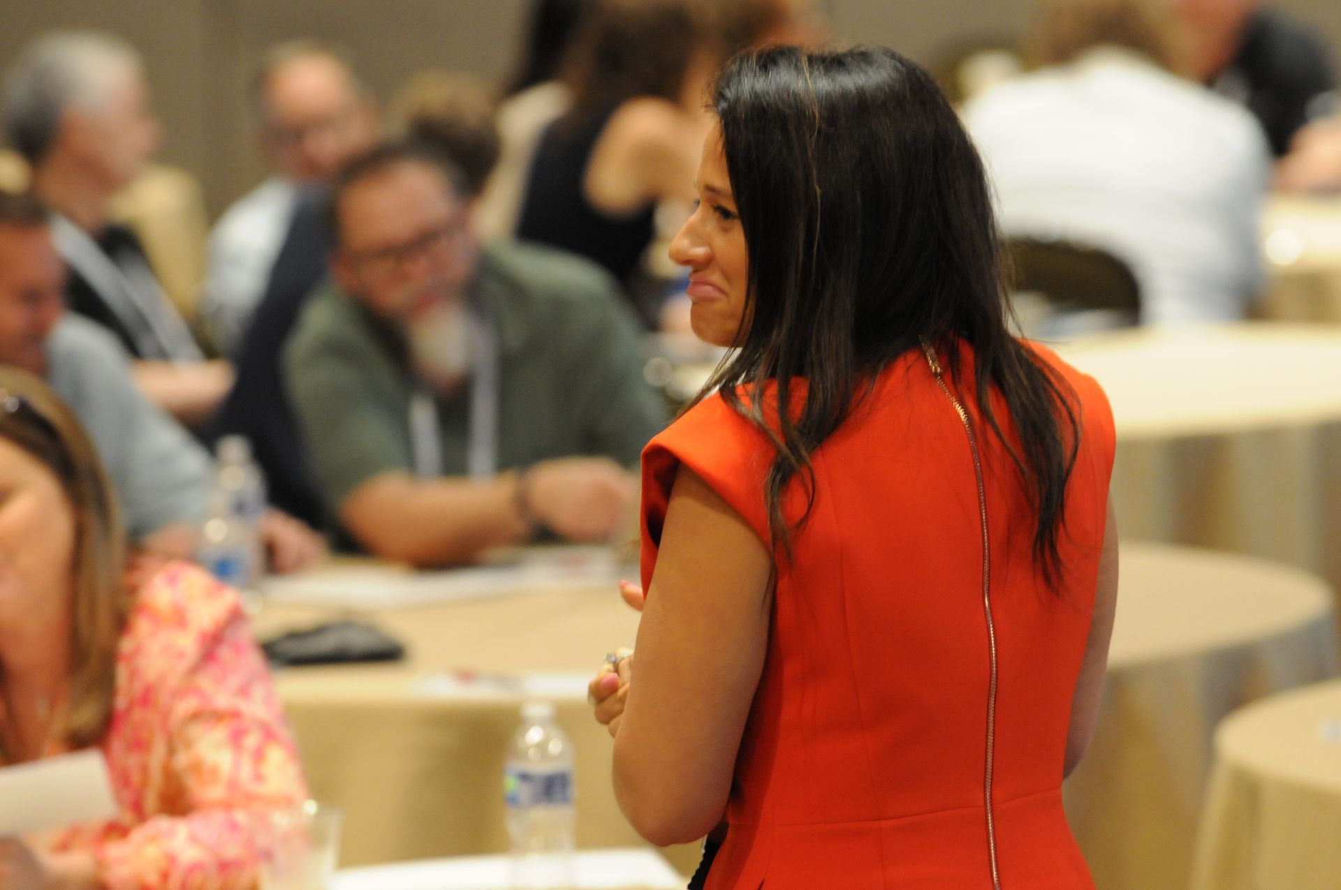 Woman in orange dress speaks at a conference. Attendees sit at round tables in a well-lit room.