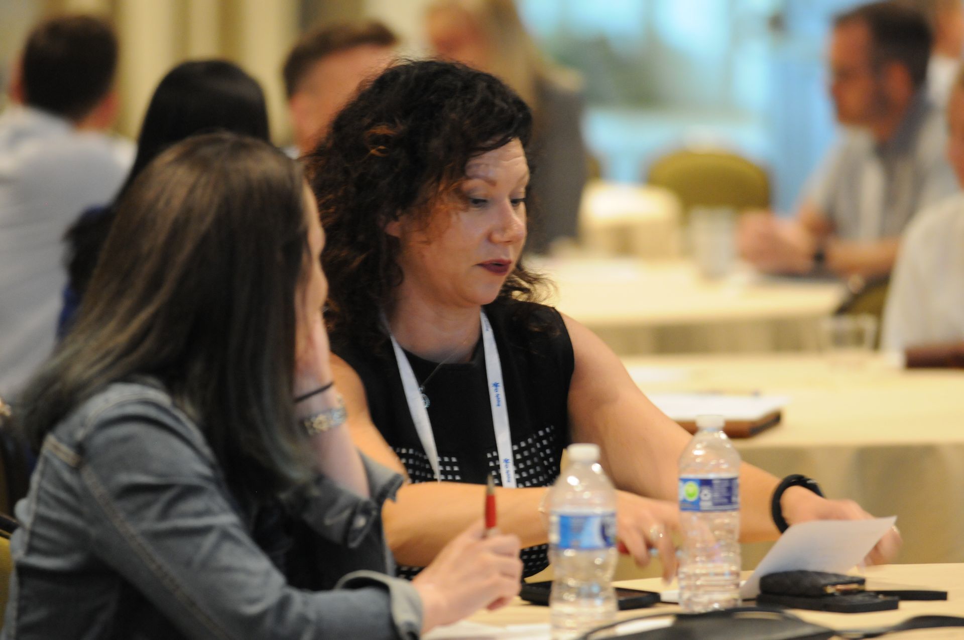 Two women at a table in a conference, talking. One has dark hair and a black top, the other has long hair and a denim jacket.