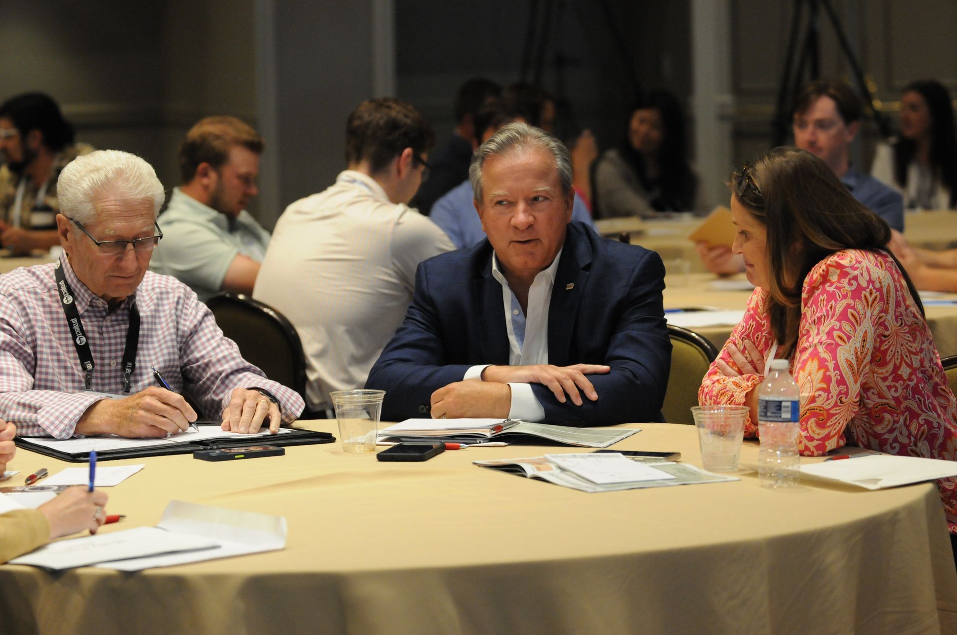 People sitting at a round table, discussing documents. Some are writing. The setting appears to be a conference.