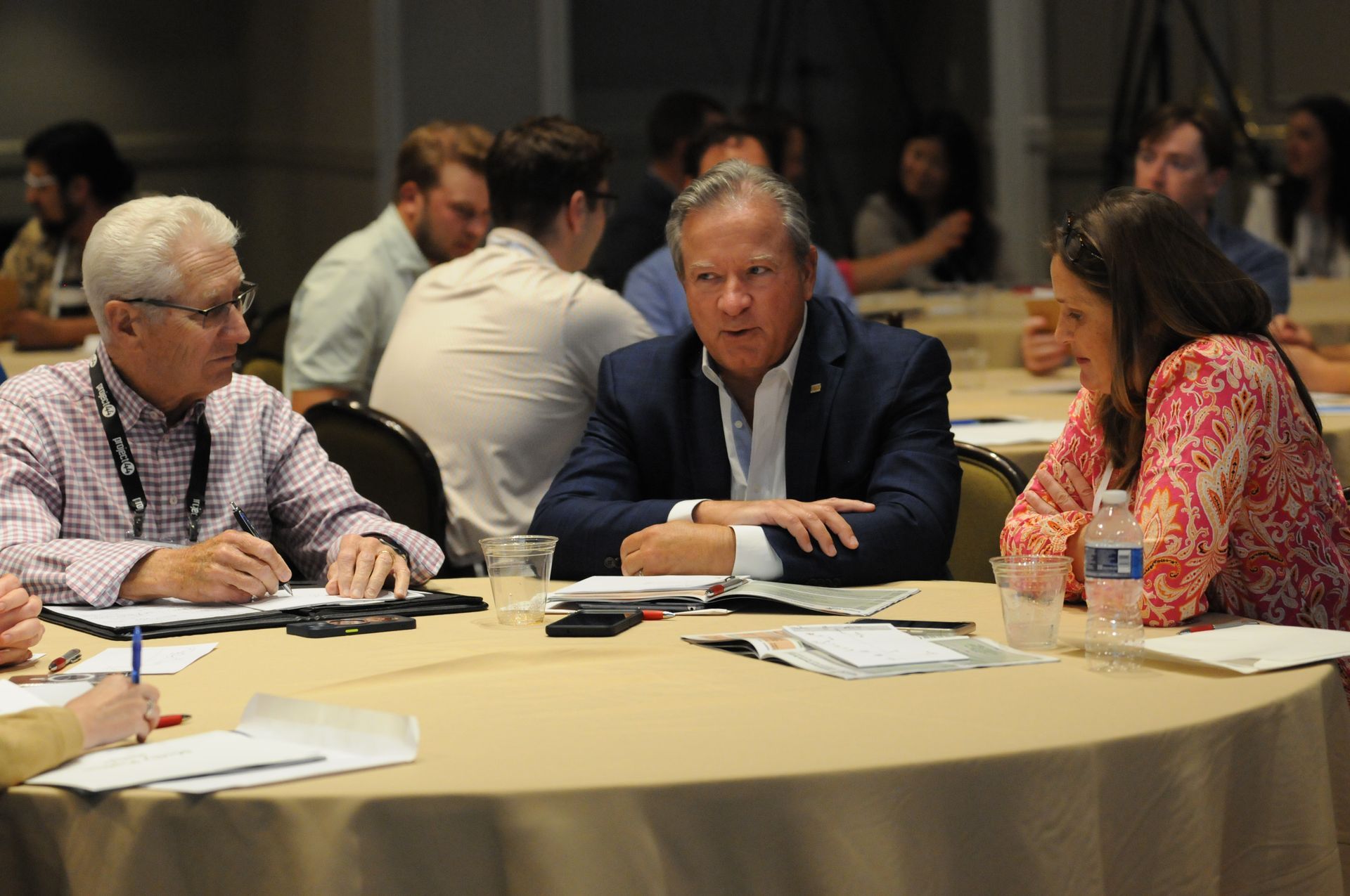 Three people at a table, discussing documents. A man in a suit and two others look on, in a conference setting.