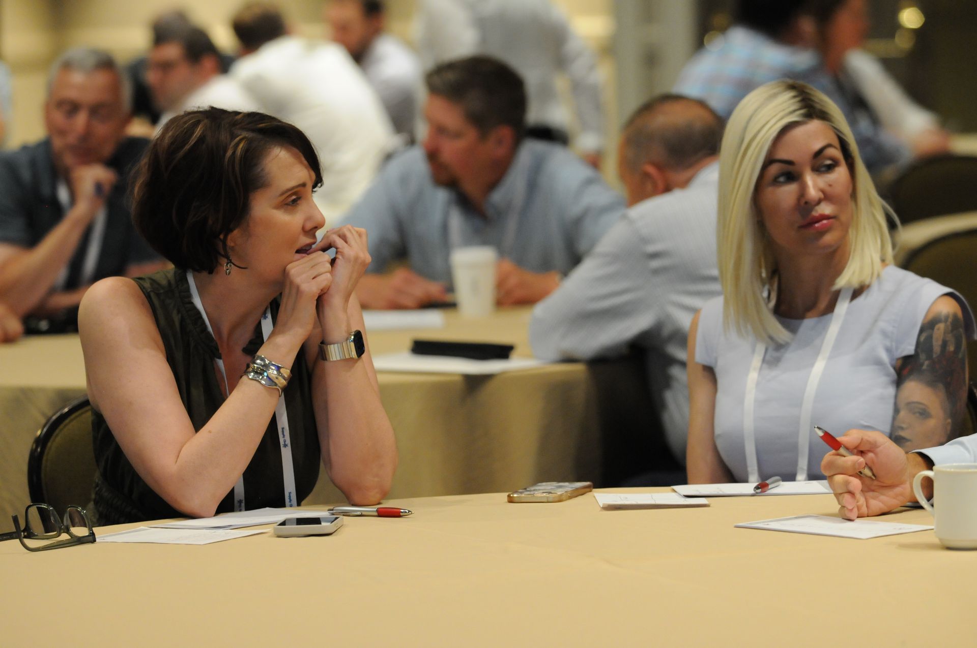 Two women at a conference table, engaged in conversation. Others in background, office setting.